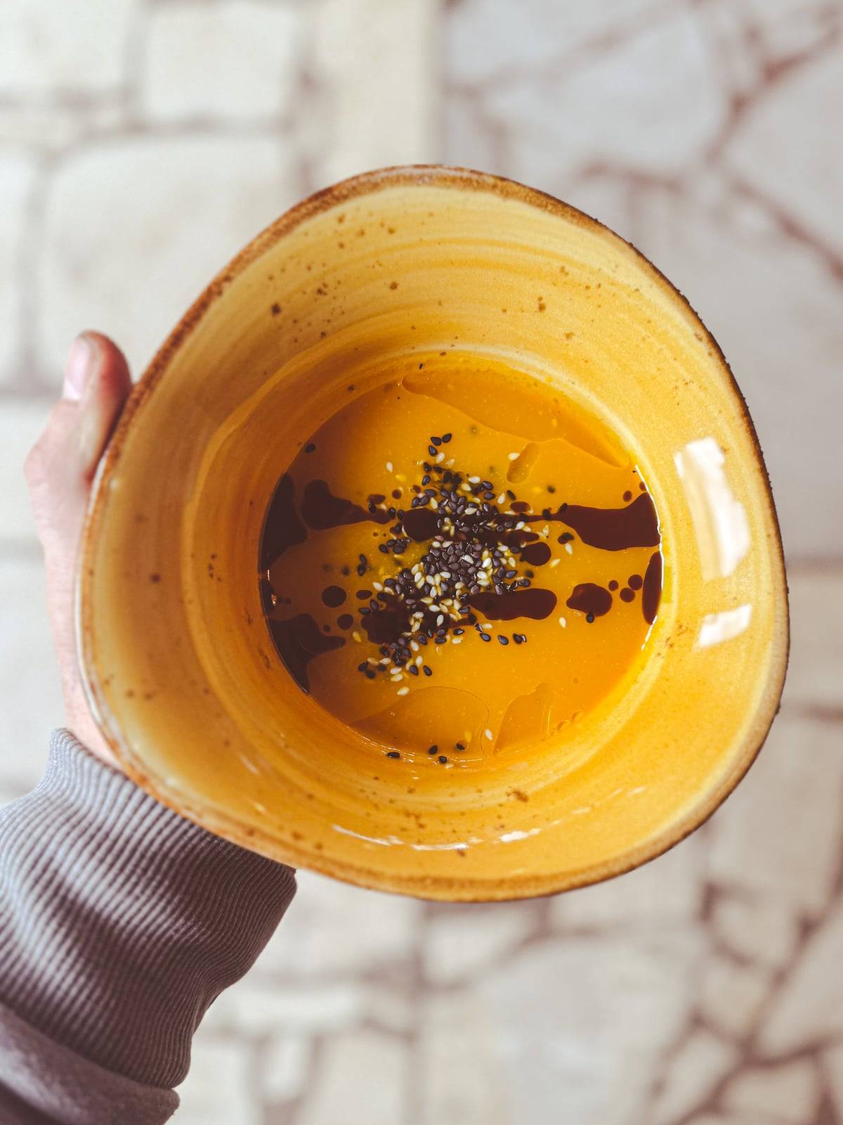 A hand holding a yellow ceramic bowl of Dr. Moro's carrot soup with sesame seeds and dark oil